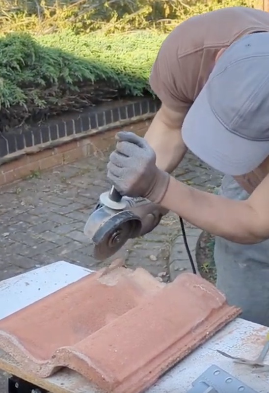 Photograph of a tile on a worktop, backside facing up. An angle grinder is visible. A person has just finished griding a channel.