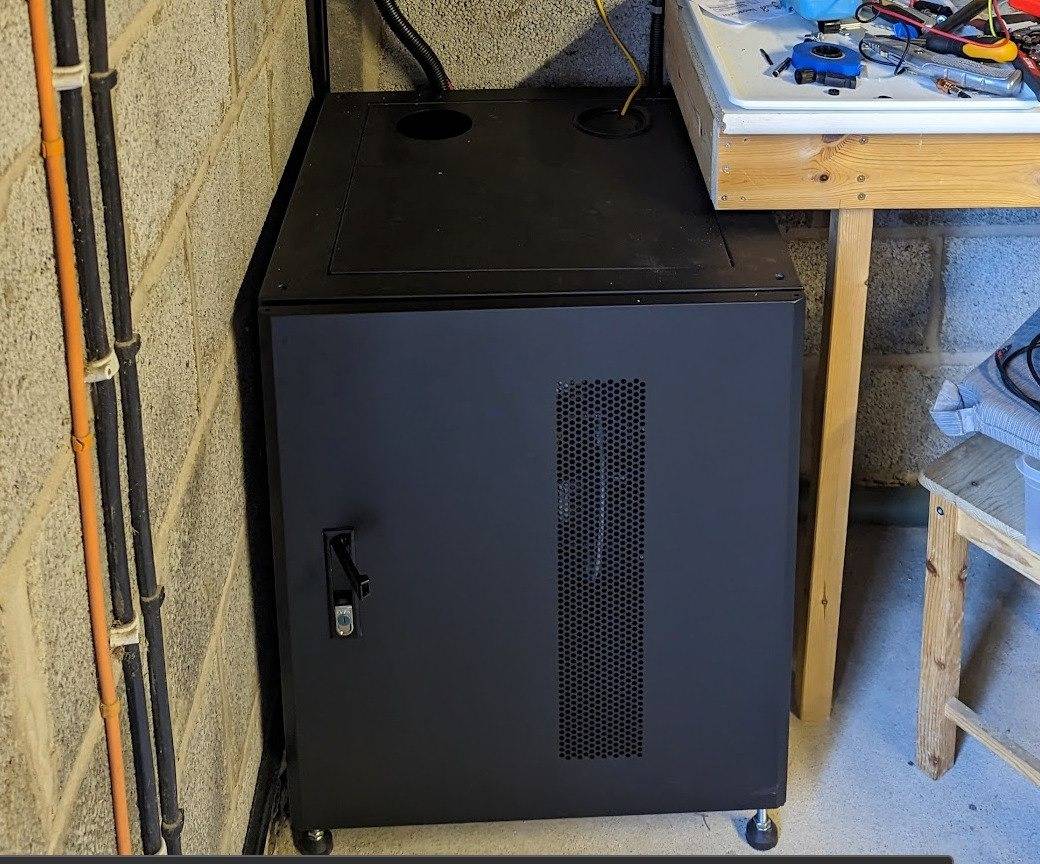 Fogstar Energy server rack cabinet installed in a garage, door closed, showing ventilation mesh, handle, lock, and cable entry holes on top