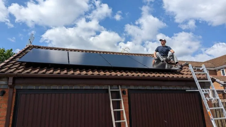A person installing solar panels on the red-tiled roof of a brick garage, with two ladders propped against the side. The person is seated on the roof under a partly cloudy blue sky in a residential neighbourhood.